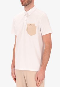 Polo bianca in cotone con una consistenza morbida, caratterizzata da una tasca color beige sul lato sinistro del petto con dettagli in bottone.