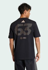 Musta puuvillainen lyhythihainen t-paita, jossa on suuri metallinhohtoinen 63-kuvio ja Mercedes-logo selässä, pyöreä pääntie, rento istuvuus.