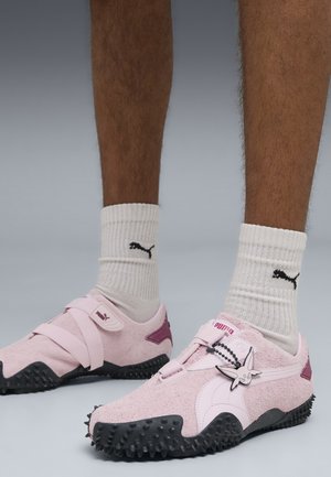 Gambe che indossano calzini bianchi Puma e sneaker rosa Puma con suole nere testurizzate e un piccolo charm a forma di cartone animato.