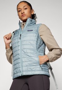 Lyseblå, quiltet vest med høy krage, glidelåslukking og sidelommer. Har en liten logo på brystet og justerbare stropper på baksiden.