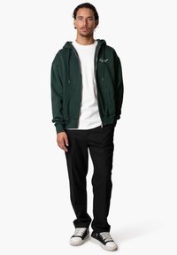 Dunkelgrüne Zip-Hoodie mit Kordelzügen, darunter ein weißes Baumwoll-T-Shirt, schwarze Hose und schwarze Sneakers mit weißen Akzenten. Einfaches Design.