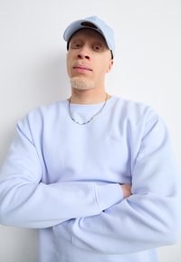Világoskék pulóver kerek nyakkal és hosszú ujjakkal, fehér hímzett logóval. Így viselve egy összeillő világoskék sapkával és ezüstlánccal.
