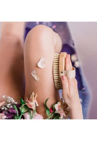 Szczotka do ciała z naturalnego włosia z drewnianą rączką trzymana przy nodze, ozdobiona płatkami kwiatów i zielenią; miękkie tekstury i neutralne kolory.