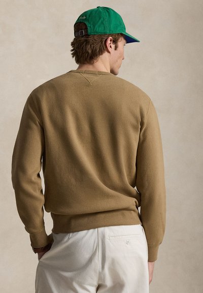 Άνδρας που φορά πράσινο καπέλο μπέιζμπολ, καφέ πουλόβερ με μακριά μανίκια και λευκό παντελόνι, στέκεται με το πίσω μέρος του σώματος προς την κάμερα μπροστά σε απλό φόντο.