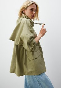 Jachetă cu glugă de culoare verde măslinie deschisă, cu mâneci largi, șnur ajustabil și buzunar frontal, asortată cu blugi de denim bleu deschis. Textură mată.