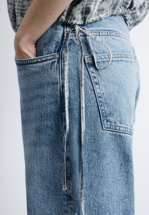 Person iført lyseblå cowboybukser med en hånd i forlommen og et mønstret stofslips hængende i siden.