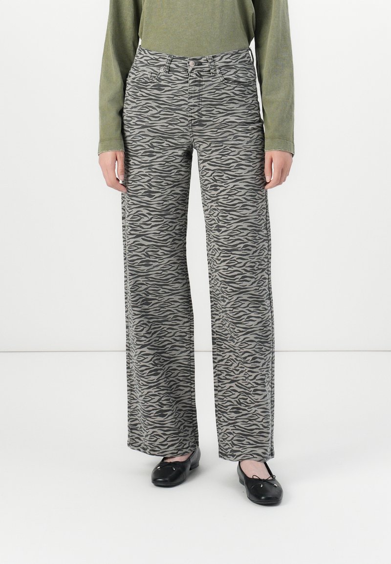 Persona che indossa pantaloni a vita alta e larghi con stampa zebra grigia e nera, un top lungo maniche verde oliva e ballerine nere.