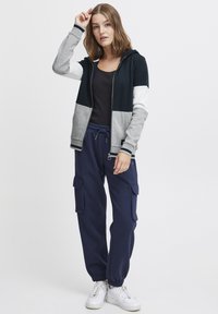 Sweatshirt à capuche coloré en bleu marine, gris et blanc ; associé à un pantalon cargo bleu marine avec poches latérales et poignets élastiques. Baskets blanches.
