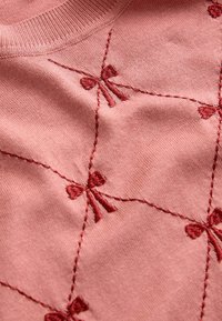 Rosa gestrickter Pullover mit bestickten Schleifen in einem Rautenmuster. Weicher Stoff mit Rundhalsausschnitt und detaillierter Naht.