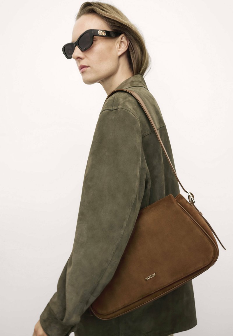 Borsa a spalla in suede marrone con forma arrotondata, tracolla regolabile e branding discreto. Il modello indossa un giaccone in suede verde oliva e occhiali da sole.