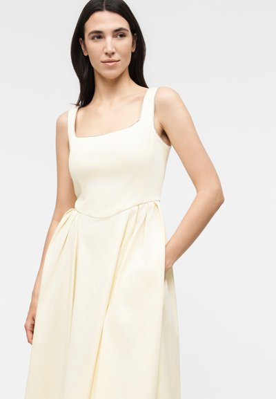 Γυναίκα με μακριά σκουρόχρωμα μαλλιά, που φοράει αμάνικο κρεμ φόρεμα με τετράγωνο ντεκολτέ και πλισέ φούστα, στέκεται με το ένα χέρι στην τσέπη.