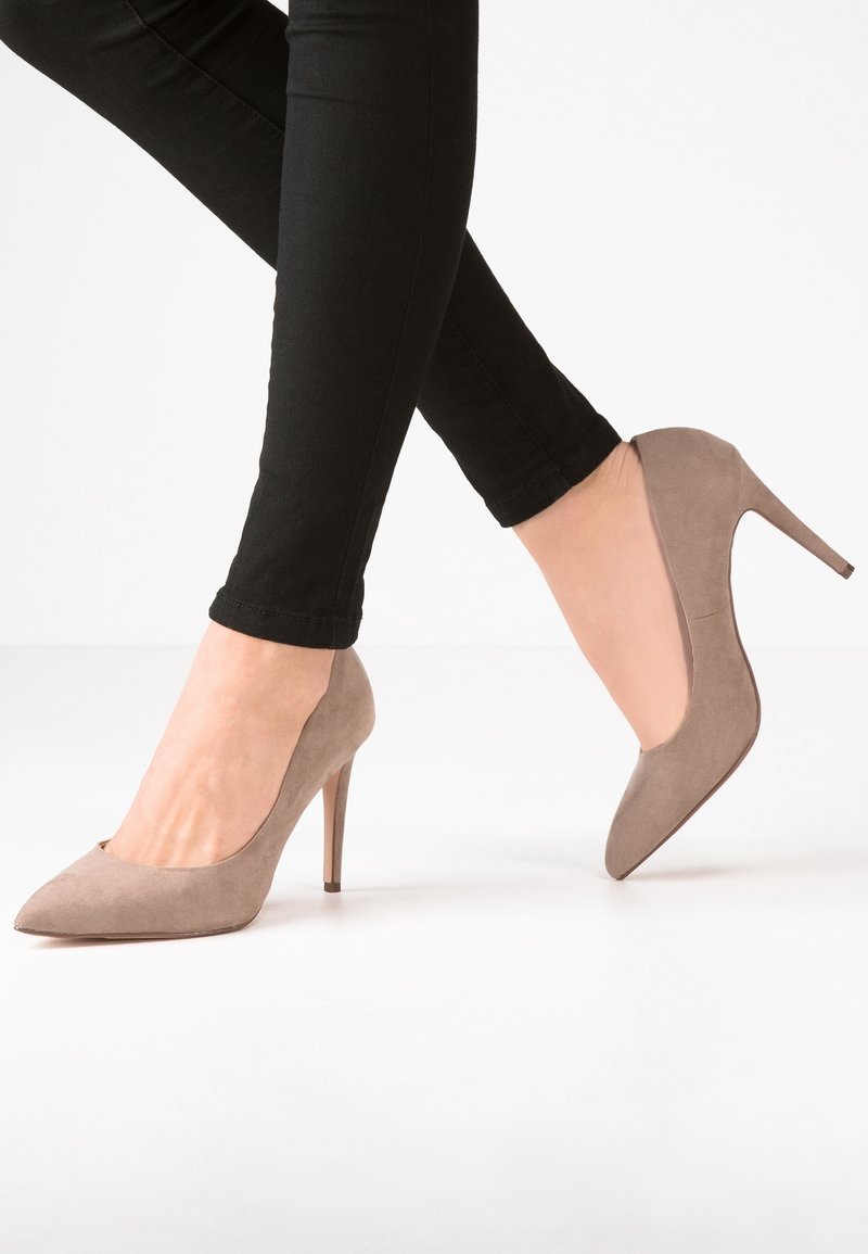 Beige mokkasini-nahkakengät korkealla korolla, teräväkärkiset ja 10 cm:n stiletto-korko, yhdistetty mustiin muotoiluun sopiviin housuihin. Sujuva pinta ja tyylikäs muotoilu.