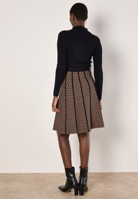Femme portant un pull noir à côtes, jupe trapèze à motifs marron et noir, bottines à talons noires, debout contre un mur uni.