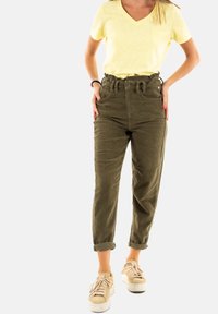 Pantalon en velours côtelé vert olive à taille haute, avec des boutons à l'avant et des poignets retroussés, associé à un t-shirt jaunt à col en v et des baskets beiges.