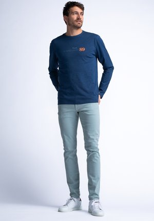 Camisa de manga larga azul marino con diseño de texto y número; combinada con unos vaqueros azul claro y zapatillas blancas, de pie contra un fondo sencillo.