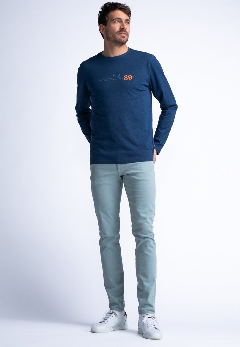 Camisa de manga larga azul marino con diseño de texto y número; combinada con unos vaqueros azul claro y zapatillas blancas, de pie contra un fondo sencillo.