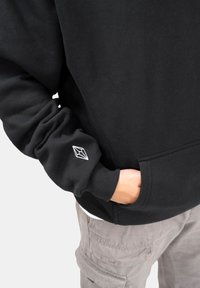 Persona con sudadera negra con un logo geométrico blanco en la manga, mano en el bolsillo delantero, pantalones cargo beige visibles abajo.