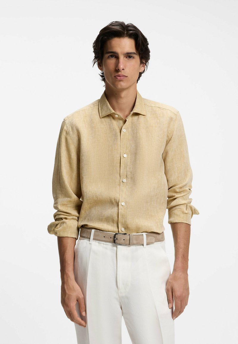 Homme portant une chemise en lin beige clair boutonnée, manches retroussées, et un pantalon blanc avec une ceinture beige sur un fond uni.