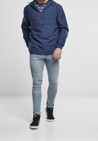 Granatowa kurtka typu pullover z ćwierć zamkiem, kieszenią z przodu i regulowanym kapturem, noszona na paskowaną koszulę i jasnoniebieskie jeansy slim fit z czarnymi sneakersami.