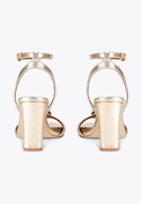 Złote metaliczne sandały na obcasie z paskiem wokół kostki, otwarty przód, grube słupkowe obcasy i gładka tekstura. Brak widocznych wzorów lub akcentów.