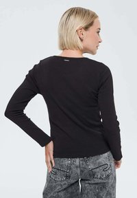 Femme aux cheveux blonds courts portant un haut à manches longues noir et un jean gris délavé, se tenant avec une main sur la hanche, faisant face à l'arrière.