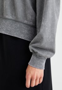 Primer plano de la mano de una persona descansando al lado de su pantalón negro, vistiendo una camiseta de manga larga gris desgastada con puños acanalados.