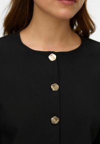 Crni odjevni predmet s okruglim izrezom na vratu i tri metalik zlatne tipke. Tkanina izgleda glatko s neupadljivim sjajem.
