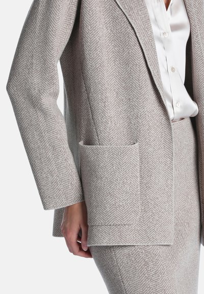 Gros plan d'une personne portant une veste de costume à chevrons gris clair et un pantalon assorti avec une chemise blanche boutonnée en dessous.