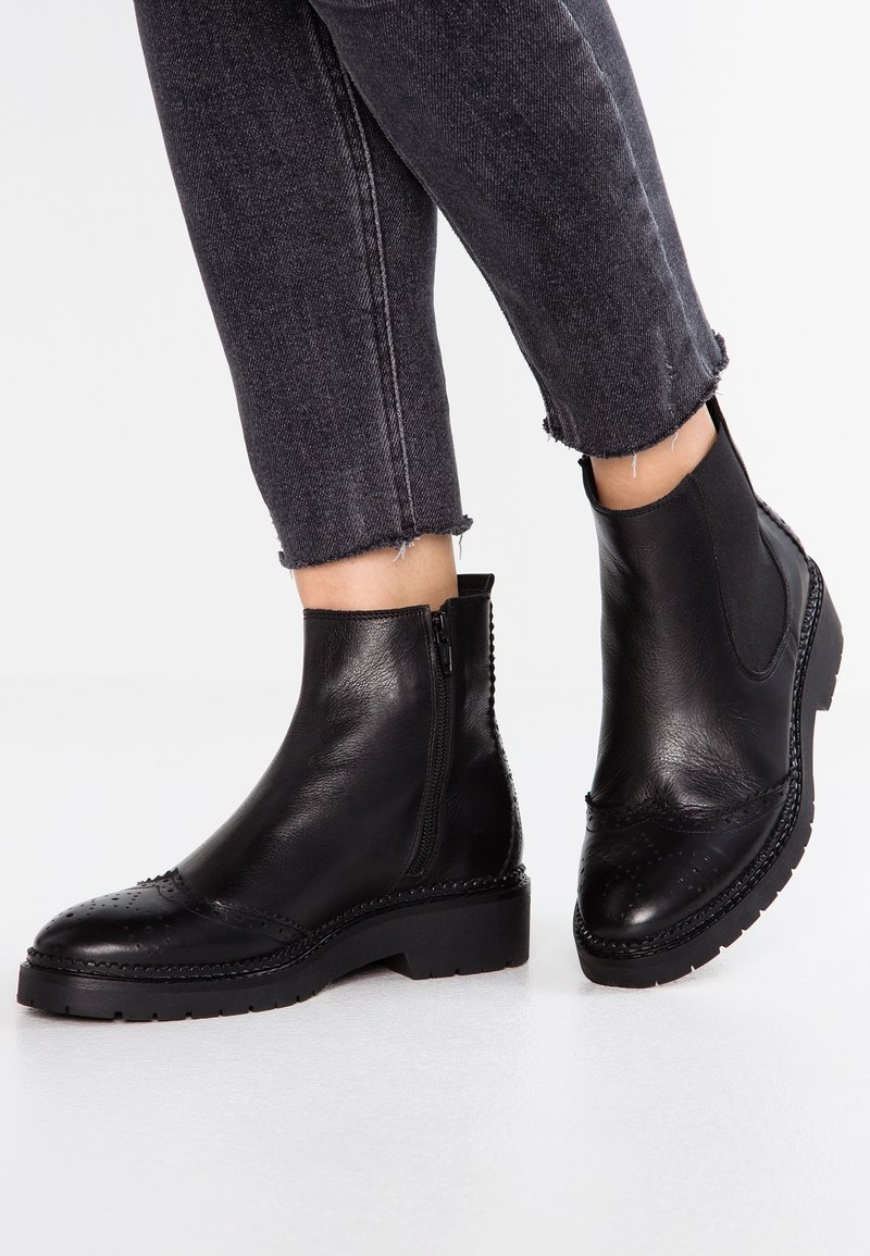 Bottes noires en cuir à la cheville avec fermetures éclair latérales portées avec un jean en denim gris foncé raccourci sur fond blanc.