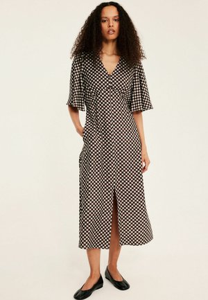 Kobieta z kręconymi włosami ubrana w czarno-białą sukienkę midi w kratę z falbanianymi rękawami i rozcięciem z przodu, stojąca z jedną ręką w kieszeni.