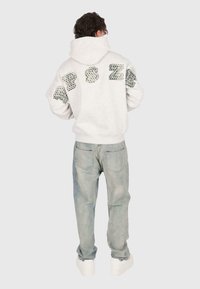 Personne de dos portant un sweat à capuche gris clair avec des lettres à motif et un jean bleu clair, ainsi que des baskets blanches sur un fond uni.