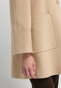 Beige wollen jas met een grote zak en textuur, gestikte manchetten. Gladde stof met een gestructureerde silhoët en rechte snit.