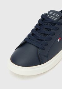 Σκούρο μπλε αθλητικό παπούτσι με λευκή σόλα, σκουρό μπλε κορδόνια και μικρό έμβλημα με κόκκινη, λευκή και μπλε σημαία στο πλάι, σε εικόνα από γωνία των δακτύλων.