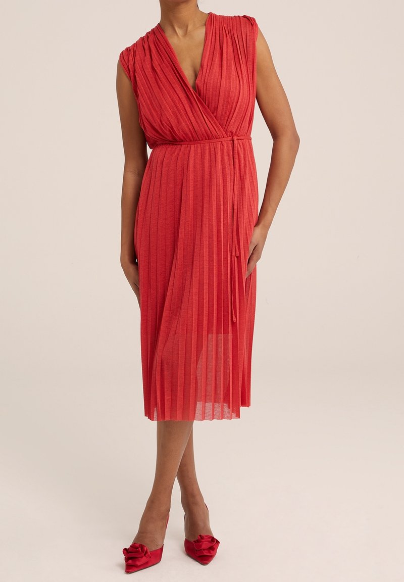 Röd plisserad klänning med en omslagdesign och djup V-ringning. Har ärmlösa axlar, knyts vid midjan och är av midi-längd. Bärs med röda klackskor.