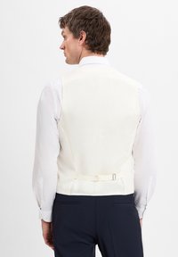 Cremfarbener Weste mit glatter Textur, die hinten mit einer kleinen Schnalle versehen ist, getragen über einem weißen Hemd, kombiniert mit dunklen Hosen.