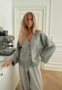 Szürke cardigan V-nyakkal, nagy ujjakra és elülső zsebekre tervezve, világosszürke, pettyezett, bő szárú nadrággal párosítva. Simább textúra, kellemes, laza dizájn.