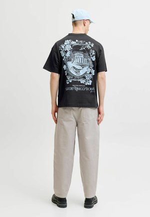 Mężczyzna w jasnoniebieskiej czapce, czarnym t-shircie z architektonicznym wzorem, beżowych spodniach i czarnych butach, stojący tyłem na białym tle.