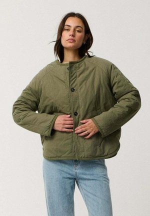 Mujer con una chaqueta acolchada de color verde oliva de gran tamaño con botones negros y vaqueros azul claro, de pie frente a un fondo liso y claro.