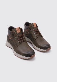 Botas de tobillo de cuero marrón con acentos grises, que presentan cordones rojos, un parche de logo color beige y una suela blanca con una suela exterior texturada en marrón.