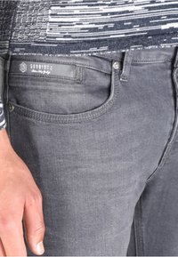 Vaqueros de mezclilla gris con un ajuste slim, que cuentan con bolsillos frontales, un botón de metal y una etiqueta con el logo en la cinturilla. Textura sutil visible.