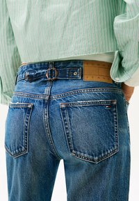 Pantalones vaqueros de denim azul con una cintura alta, que cuentan con bolsillos traseros, una etiqueta de cuero marrón con el logo "TOMMY JEANS" y costuras en contraste.