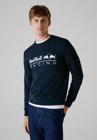 Sudadera azul marino de manga larga, con diseño de cuello redondo, que presenta un prominente logo blanco de "Red Bull Racing" y dos gráficos de toros en posición de carga.