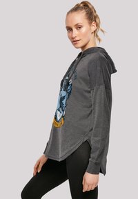 Grå hettegenser med rund kant, med blå og gul Ravenclaw-emblem. Lange ermer og myk tekstur.
