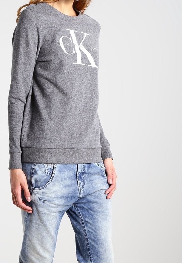 Un sweat-shirt gris avec un logo "CK" blanc sur le devant, doté d'un col rond et de poignets côtelés, associé à un jean en denim bleu clair.