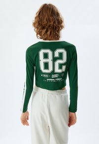 Vihreä pitkähihaisuus crop-top, jossa on valkoisia yksityiskohtia, takana numero "82" ja teksti, valmistettu sileästä, venyvästä kankaasta.