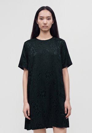 Femme aux longs cheveux noirs portant une robe noire à manches courtes avec une texture florale, debout devant un fond clair uni.