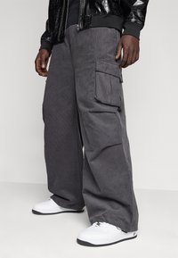 Šedé pracovní kalhoty z manšestru s velkými bočními kapsami, volným střihem, doplněné bílými teniskami. Detail texturované tkaniny je viditelný.