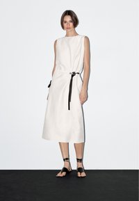 Weißes, ärmelloses Leinenkleid mit Gürtel in der Taille, gerader Schnitt und seitlichen Taschen. Kombiniert mit schwarzen Riemchensandalen.