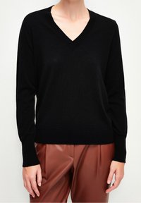 Svart stickad tröja med V-ringning och långa ärmar, med en mjuk textur. Matchas med bruna veckade byxor.