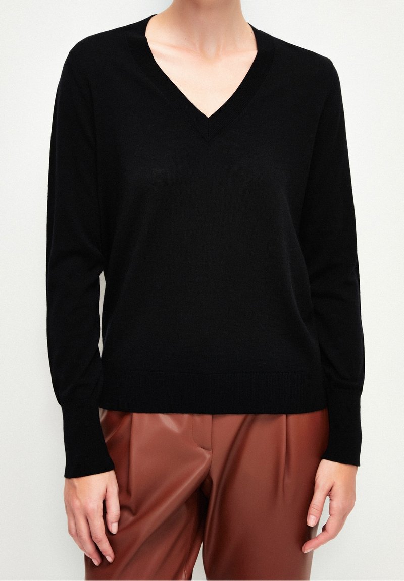 Svart stickad tröja med V-ringning och långa ärmar, med en mjuk textur. Matchas med bruna veckade byxor.